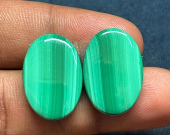 May include: Two oval-shaped malachite gemstones. The polished stones display a vibrant green color with distinctive banded patterns. The gemstones are held between fingers, showcasing their smooth, glossy surfaces. The stones are approximately 1 inch in length.