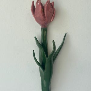 Könnte beinhalten: Eine Wandskulptur aus Keramik in Form einer Tulpenblüte. Die Blüte ist in einem staubigen Rosaton mit glänzender Oberfläche gehalten, während der Stiel und die Blätter in einem tiefen, kräftigen Grün mit glatter, reflektierender Glasur gestaltet sind.