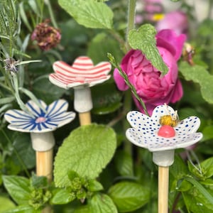 Puede incluir: Tres estacas de jardín de flores de cerámica con diseños blancos, rojos y azules. Las estacas están en un jardín con follaje verde y una rosa rosa en el fondo. Las estacas de flores tienen forma de hongo.