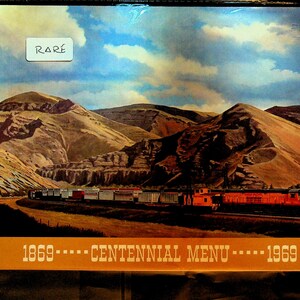 May include: A rectangular menu cover featuring a landscape painting of a freight train traveling through a mountainous region under a blue sky with clouds. The text "1869 ---- CENTENNIAL MENU ---- 1969" is printed in white on an orange band.