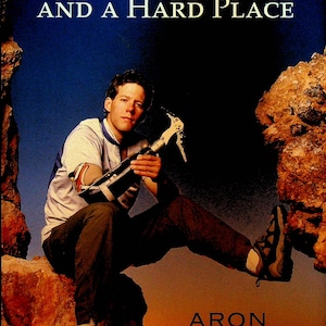 May include: A book cover featuring a man sitting on a rock with a blue sky and a sunset in the background. The book title is "Between a Rock and a Hard Place" and the author is "Aron Ralston".