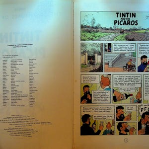 May include: Open book with comic strip panels from "Tintin and the Picaros." The title is displayed at the top. The left page lists the countries where the book was published. The comic panels depict scenes with characters and speech bubbles.