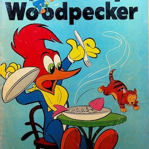 May include: Vintage comic book cover featuring Woody Woodpecker. Woody, with his red head and blue outfit, is seated at a table with a plate of fish. A cat leaps away. The title "Woody Woodpecker" is in large black letters.