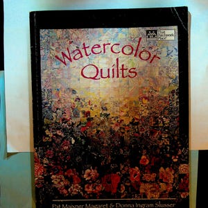 Puede incluir: Un libro titulado "Watercolor Quilts" con una cubierta negra. La portada presenta un diseño de patchwork colorido de patrones florales. El título está en escritura roja, y los nombres de los autores están en la parte inferior.