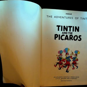 May include: Open book featuring the cover of "Tintin and the Picaros." The title is in bold black letters. Below the title is a colorful illustration of three figures in red and blue outfits. The book is published by Little, Brown and Company.