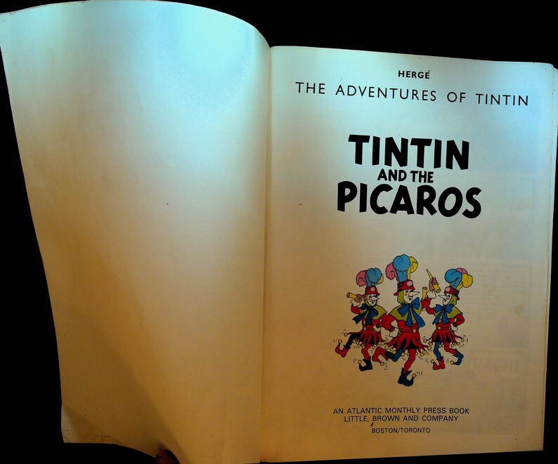 May include: Open book featuring the cover of "Tintin and the Picaros." The title is in bold black letters. Below the title is a colorful illustration of three figures in red and blue outfits. The book is published by Little, Brown and Company.