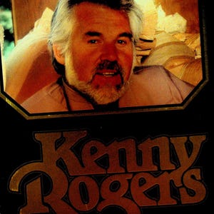 Op de afbeelding: Een ingelijst portret van Kenny Rogers. De afbeelding toont een man met grijs haar en een baard, gekleed in een lichtgekleurd overhemd. De naam "Kenny Rogers" wordt weergegeven in een gestileerd, oranje lettertype tegen een zwarte achtergrond.