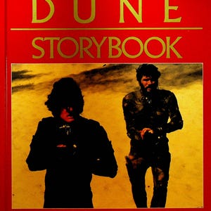 May include: The Dune Storybook cover features a red background with gold lettering. The title "THE DUNE STORYBOOK" is displayed in large, bold letters. The cover also includes a black and white photo of two figures in a desert setting.