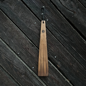 May include: A wooden shoehorn with a black cord loop at the top. The shoehorn is hanging on a dark wood wall.