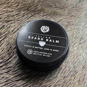 May include: A black tin container with a white label that reads "TALLOW BEARD BALM" and "SIMPLE IS BETTER. LESS IS MORE."  The label also features a bear head logo and a beard icon. The container is labeled "BEYOND REWILDFARMS.COM SEATTLE, WA USA".