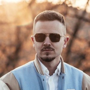 Op de afbeelding: Een man draagt een blauw en bruin varsity jack met een wit overhemd met kraag eronder. Hij draagt een zonnebril met een gouden montuur.