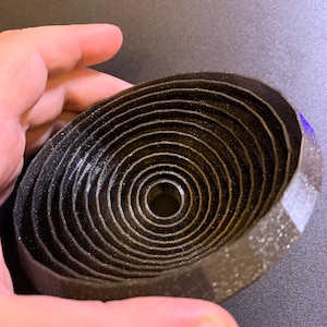 May include: A black, circular, 3D printed object with concentric rings. The object is made of a textured material and has a small hole in the center.