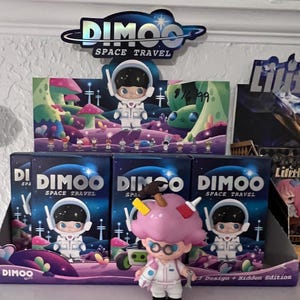 May include: A display of DIMOO Space Travel figurines. The boxes and figurines feature a space theme with a cartoon astronaut. One figurine has a pink ice cream cone-shaped head. The boxes and sign read "DIMOO SPACE TRAVEL".