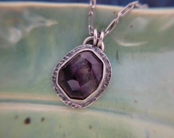May include: A pendant necklace featuring a faceted, deep purple gemstone set in a textured, hammered silver frame. The pendant hangs from a silver chain with elongated oval links, showcasing a dark, irregular shape and a polished metallic finish.