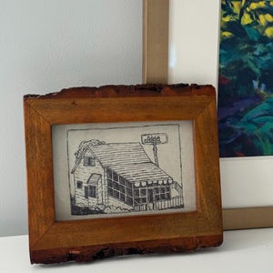 May include: A framed black and white line drawing of a house with a sign that reads "Adele's". The frame is made of wood with a natural bark edge.