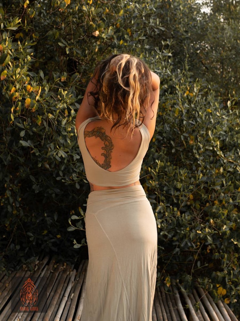 Puede incluir: Un conjunto de dos piezas verde oliva claro, que consta de un top corto y una falda larga. La modelo est&aacute; de pie frente a un fondo de hojas verdes. Un tatuaje es visible en la espalda de la modelo. El logotipo "CARIK GANG" est&aacute; en la esquina inferior izquierda.