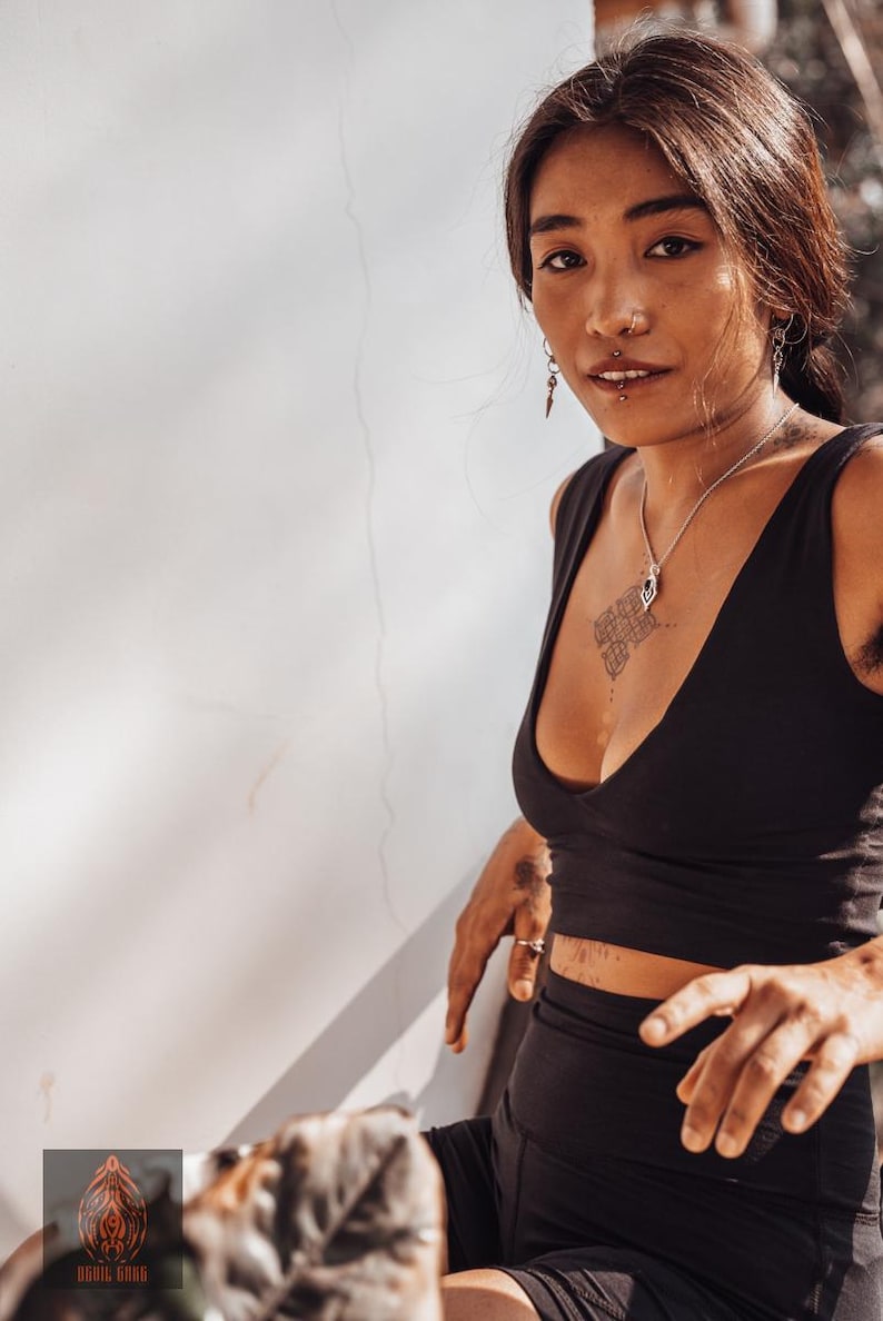 Puede incluir: Una mujer con un top corto y pantalones cortos negros. Tiene un collar y pendientes de plata. La mujer tiene tatuajes y un piercing en la nariz. La imagen est&aacute; tomada con luz natural.