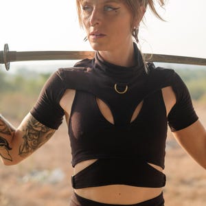 Könnte beinhalten: Schwarzes Crop-Top mit hohem Ausschnitt und Cut-Out-Design, mit goldenem Ringdetail. Das Top wird mit passenden schwarzen Hosen kombiniert. Ein Schwert ruht auf den Schultern.