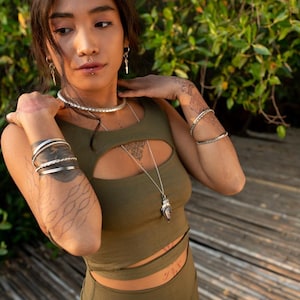 Puede incluir: Conjunto de top corto y pantal&oacute;n verde oliva. El top tiene un corte de ojo de cerradura y se combina con un collar largo de plata. La modelo lleva varias pulseras de plata y una gargantilla de plata. Se ven tatuajes en los brazos.