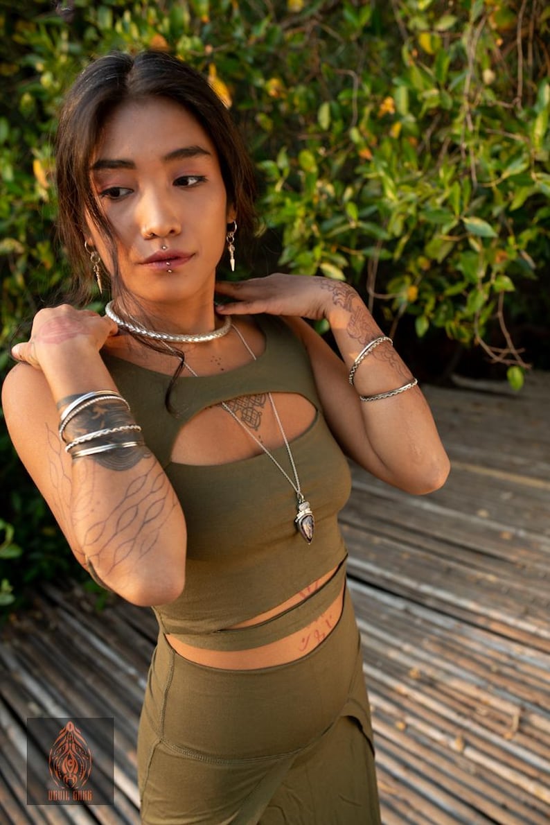 Puede incluir: Conjunto de top corto y pantal&oacute;n verde oliva. El top tiene un corte de ojo de cerradura y se combina con un collar largo de plata. La modelo lleva varias pulseras de plata y una gargantilla de plata. Se ven tatuajes en los brazos.
