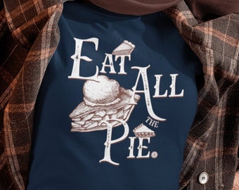 May include: A navy blue t-shirt with a white graphic that says "Eat All The Pie." The shirt is being worn by a person lying on a bed of brown leaves.