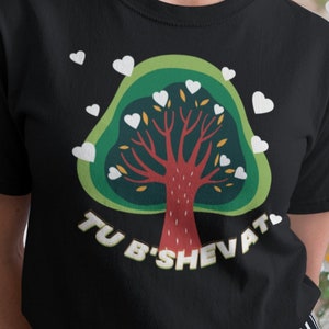 May include: Black t-shirt featuring a tree design with a red trunk, green leaves, and white heart-shaped accents. The text "TU B'SHEVAT" is printed below the tree. The person is wearing black and white striped pants.