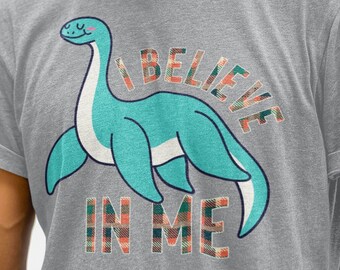 May include: Sport grey heather t-shirt featuring a teal dinosaur illustration with the text "I BELIEVE IN ME" in a plaid pattern. The shirt is a crew neck and has short sleeves. The text "sport grey heather" is at the bottom.