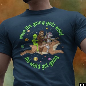 May include: Navy blue t-shirt featuring a whimsical design of an alien, Bigfoot, and a unicorn riding a jackalope. The text "when the going gets weird, the weird get going" is printed in green around the image.