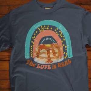 Puede incluir: Camiseta azul marino con un gr&aacute;fico de arco&iacute;ris y una pila de panqueques con jarabe. El texto "The Love is Real" est&aacute; impreso en la camiseta.