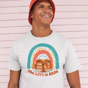 Puede incluir: Una camiseta blanca con un gr&aacute;fico de arco&iacute;ris y el texto "The LOVE is REAL". El gr&aacute;fico presenta una pila de panqueques con jarabe y mantequilla. El modelo lleva un sombrero de pescador rojo.