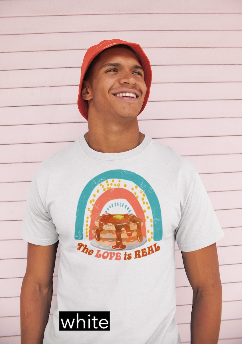 Puede incluir: Una camiseta blanca con un gr&aacute;fico de arco&iacute;ris y el texto "The LOVE is REAL". El gr&aacute;fico presenta una pila de panqueques con jarabe y mantequilla. El modelo lleva un sombrero de pescador rojo.