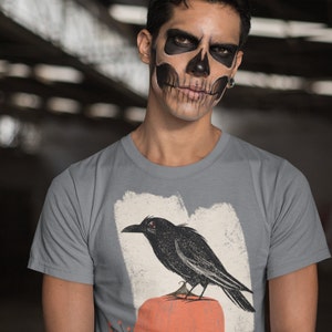 May include: A gray t-shirt with a black crow perched on a pumpkin. The crow has red eyes and the pumpkin is orange. The design is on a white background with a distressed look.