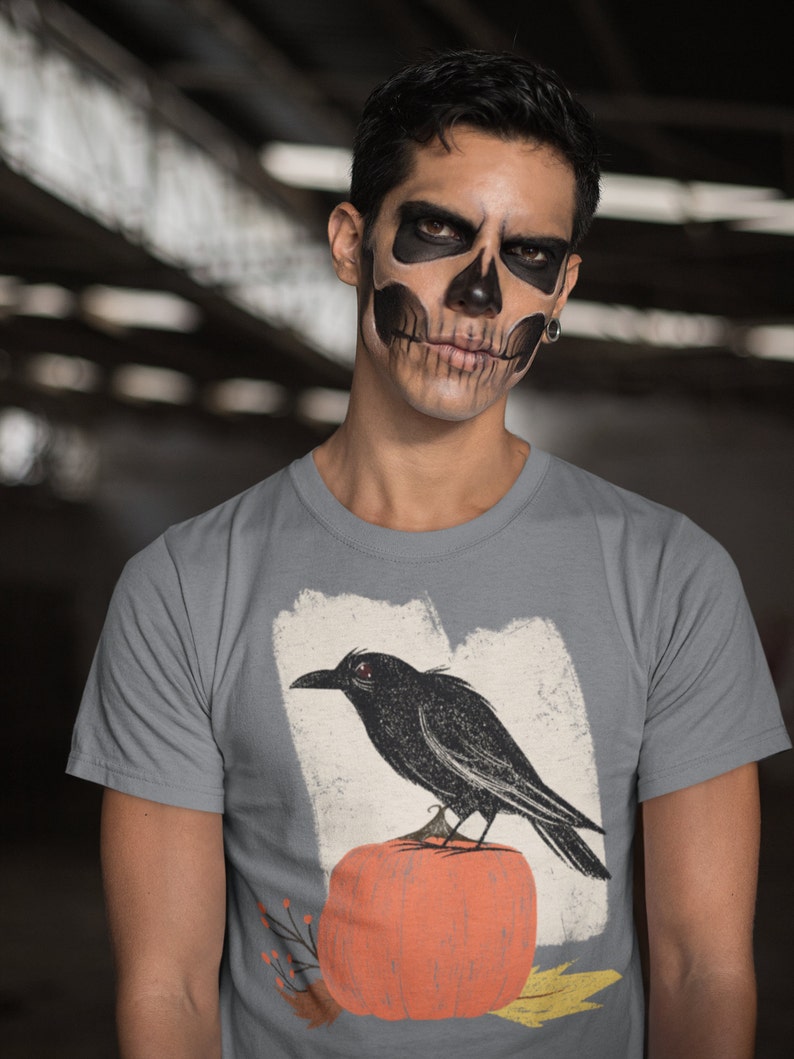 May include: A gray t-shirt with a black crow perched on a pumpkin. The crow has red eyes and the pumpkin is orange. The design is on a white background with a distressed look.
