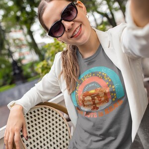 Puede incluir: Una camiseta gris antracita con un gr&aacute;fico de arco&iacute;ris de estilo vintage y el texto "The Love is Real" con una pila de panqueques en el centro del arco&iacute;ris.