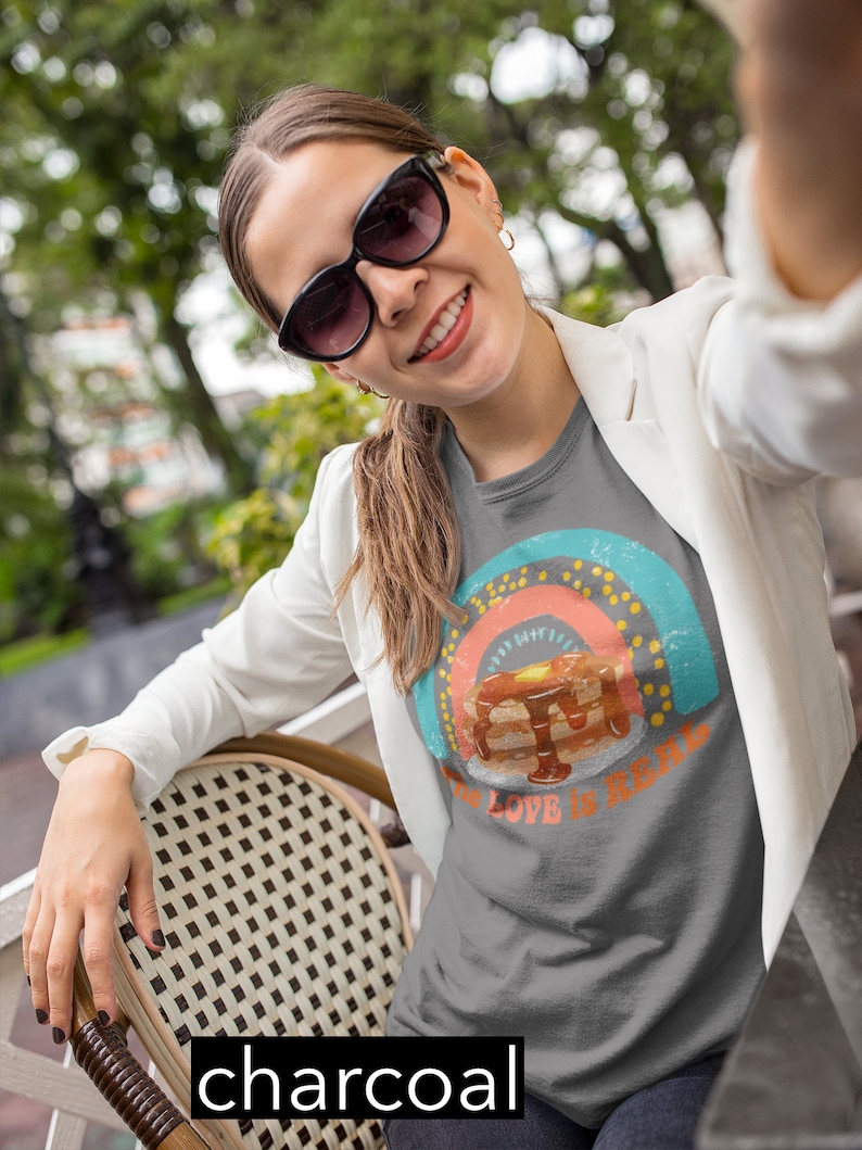 Puede incluir: Una camiseta gris antracita con un gr&aacute;fico de arco&iacute;ris de estilo vintage y el texto "The Love is Real" con una pila de panqueques en el centro del arco&iacute;ris.