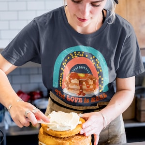 Puede incluir: Una persona lleva una camiseta azul marino con un gr&aacute;fico de arco&iacute;ris y panqueques que dice "The Love is Real". Est&aacute; glaseando un pastel de tres pisos con glaseado blanco.