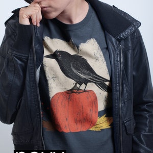 May include: A navy blue t-shirt with a graphic of a black crow perched on a red pumpkin. The t-shirt is worn by a person wearing a black leather jacket.
