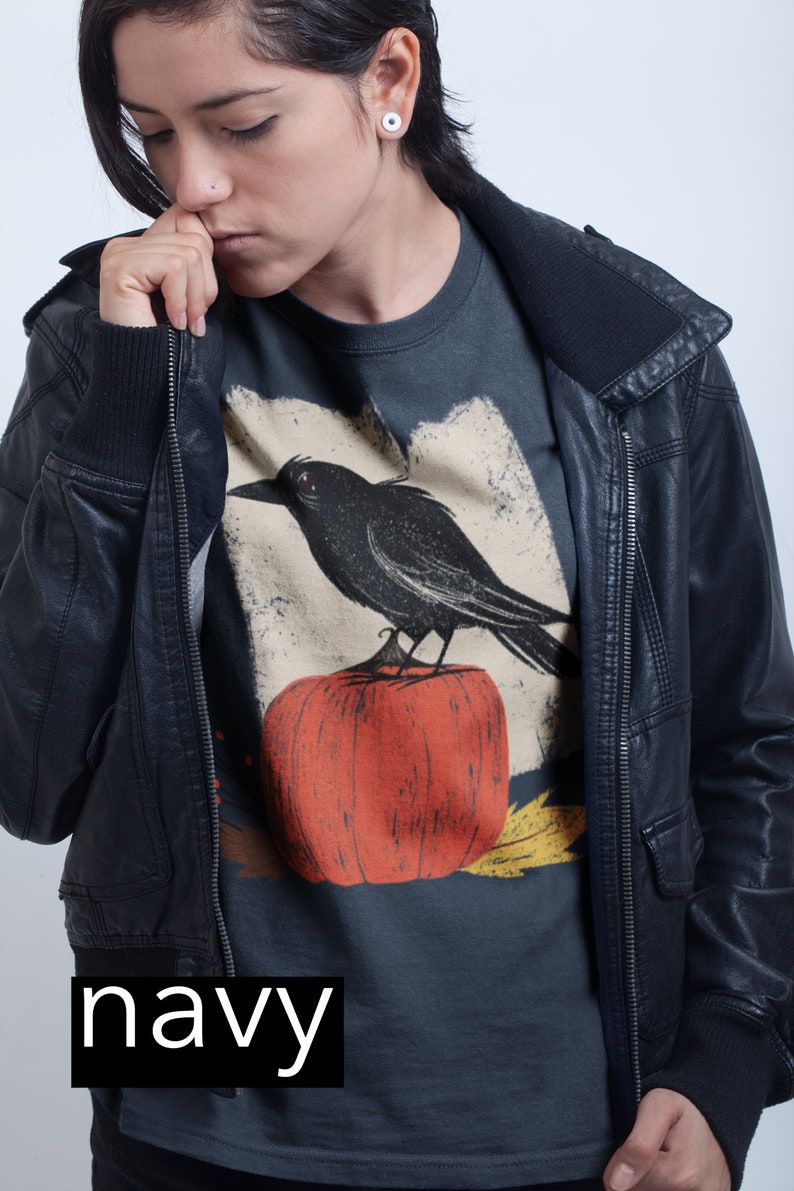 May include: A navy blue t-shirt with a graphic of a black crow perched on a red pumpkin. The t-shirt is worn by a person wearing a black leather jacket.