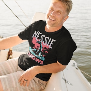 May include: A man wearing a black t-shirt with a graphic of a sea monster and the text "Nessie Knows" in pink and blue lettering. He is sitting on a boat and smiling.