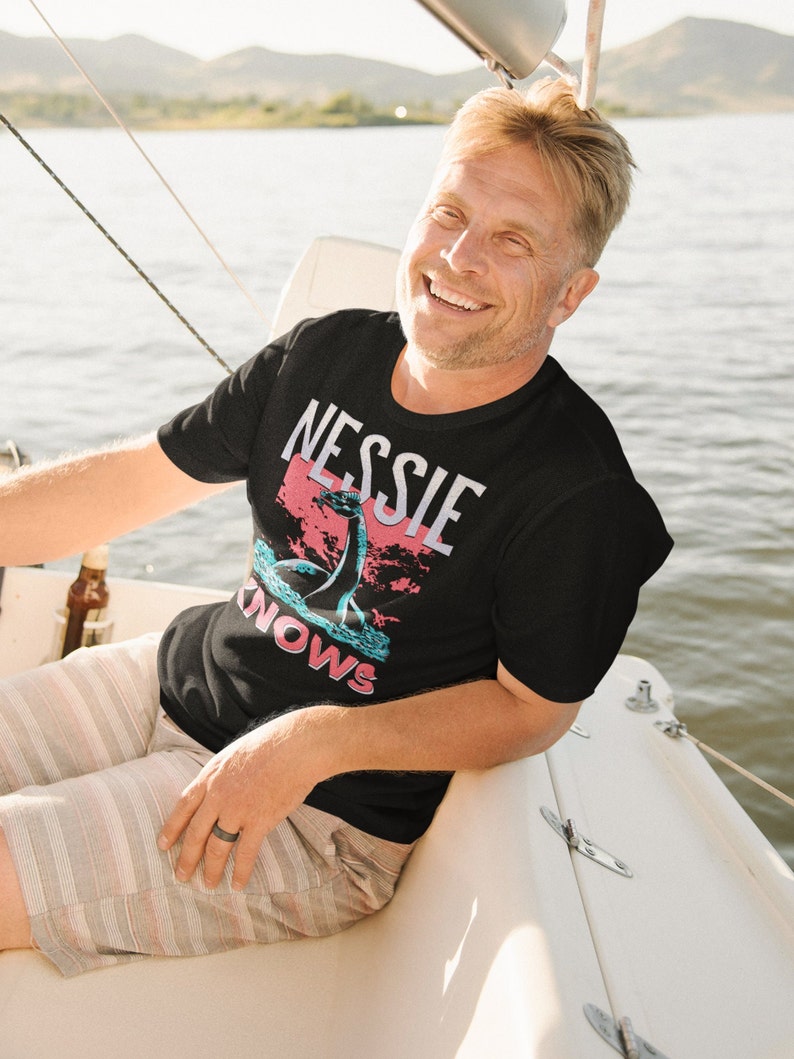 May include: A man wearing a black t-shirt with a graphic of a sea monster and the text "Nessie Knows" in pink and blue lettering. He is sitting on a boat and smiling.