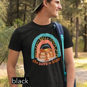 Puede incluir: Una camiseta negra con un gr&aacute;fico de arco&iacute;ris y el texto "The LOVE is REAL". El gr&aacute;fico presenta una pila de panqueques con jarabe y una porci&oacute;n de mantequilla.
