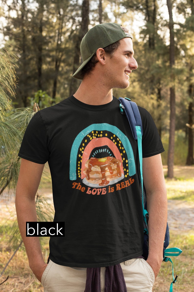 Puede incluir: Una camiseta negra con un gr&aacute;fico de arco&iacute;ris y el texto "The LOVE is REAL". El gr&aacute;fico presenta una pila de panqueques con jarabe y una porci&oacute;n de mantequilla.