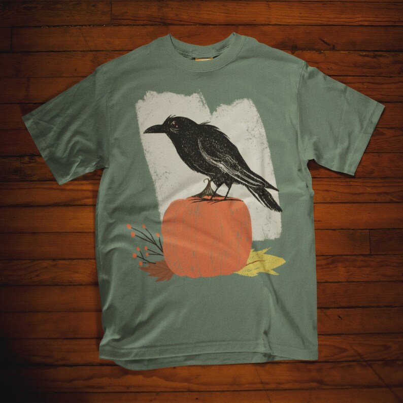 May include: A teal blue t-shirt with a black crow perched on top of an orange pumpkin. The crow is facing the left side of the image. The pumpkin is surrounded by small brown branches and leaves.