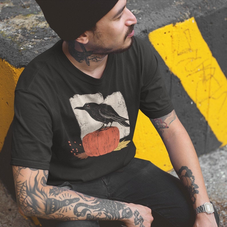 May include: A black t-shirt with a vintage-style illustration of a black crow perched on top of an orange pumpkin. The illustration is surrounded by a white border and has a distressed, faded look.