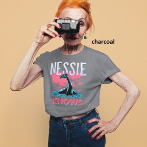 May include: A charcoal grey t-shirt with a graphic of a Loch Ness Monster and the text "Nessie Knows".