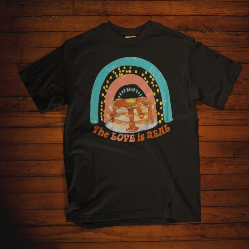 Puede incluir: Camiseta negra con un gr&aacute;fico de arco&iacute;ris y una pila de panqueques con jarabe. El texto "The LOVE is REAL" est&aacute; impreso debajo del gr&aacute;fico.