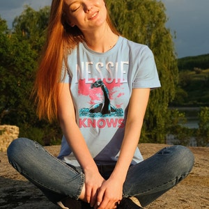 May include: A woman wearing a light blue t-shirt with a graphic of a Loch Ness Monster and the text "Nessie Knows". She is sitting with her legs crossed and wearing blue jeans and black sneakers.