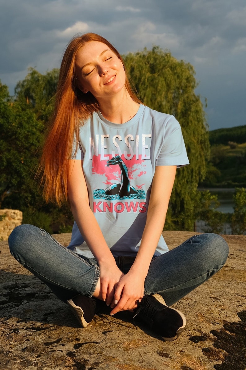 May include: A woman wearing a light blue t-shirt with a graphic of a Loch Ness Monster and the text "Nessie Knows". She is sitting with her legs crossed and wearing blue jeans and black sneakers.