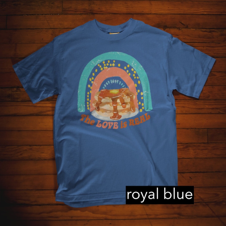 Puede incluir: Una camiseta azul real con un gr&aacute;fico de arco&iacute;ris de estilo vintage y una pila de panqueques con jarabe. El texto "The Love is Real" est&aacute; impreso debajo de los panqueques.
