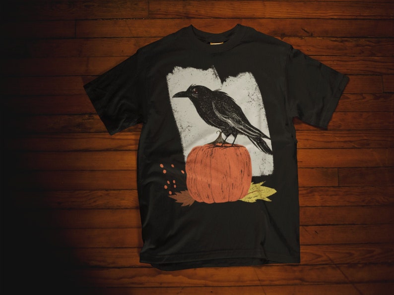 May include: Black t-shirt with a graphic print of a crow perched on a pumpkin. The pumpkin is orange and the crow is black with a white background.